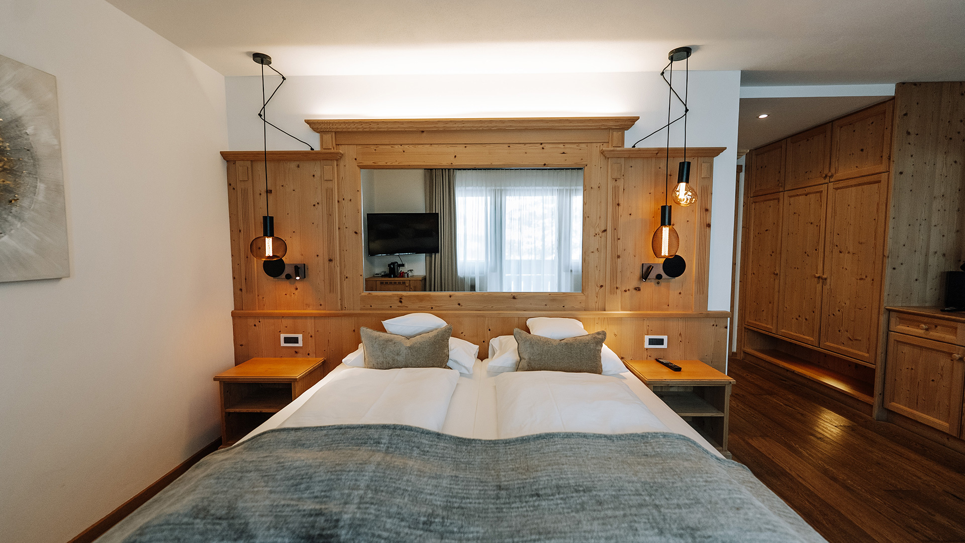 Alpines Zimmer mit hellen Holzmöbeln, Kopfteil mit integriertem Spiegel, Hängelampen und offenem Kleiderschrank.