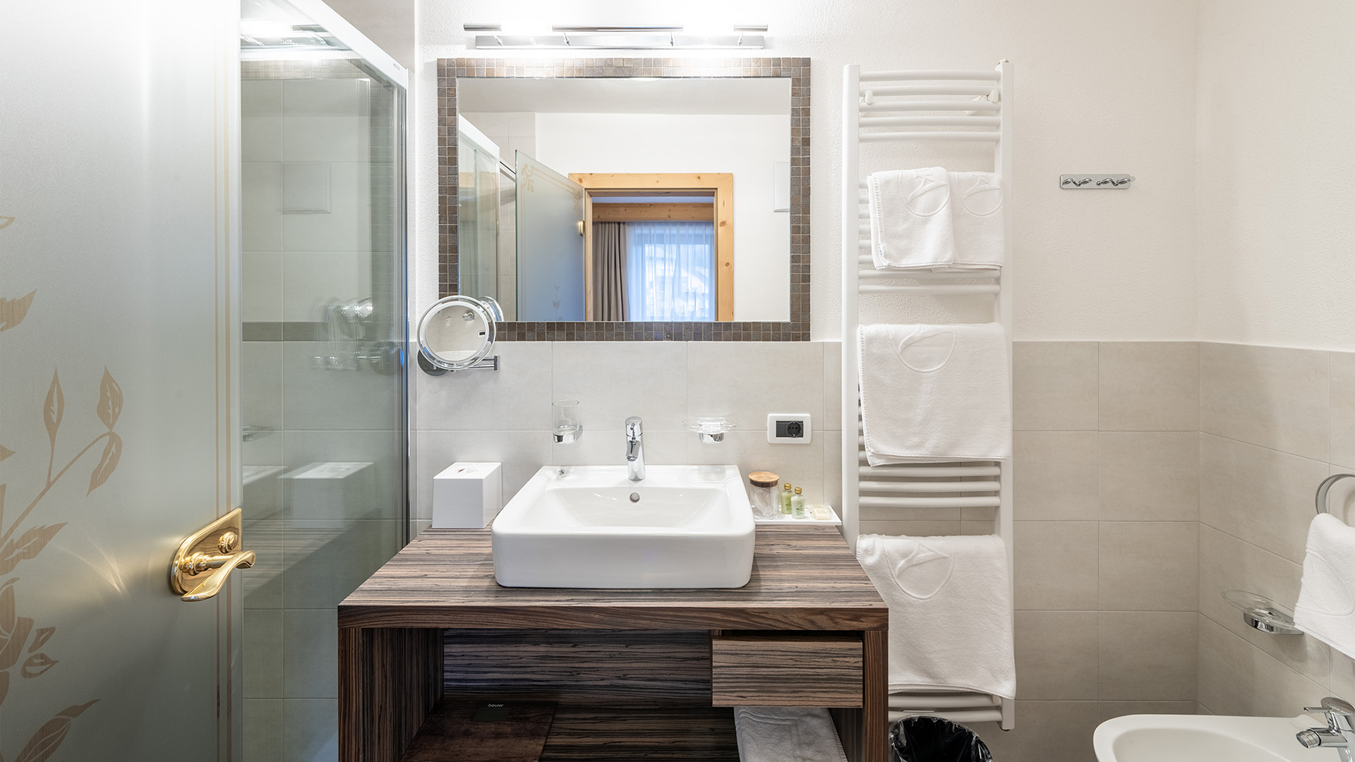 Modernes Badezimmer mit Waschbecken auf Holzplatte, beleuchtetem Spiegel, Glasdusche und Handtuchheizkörper mit Handtüchern