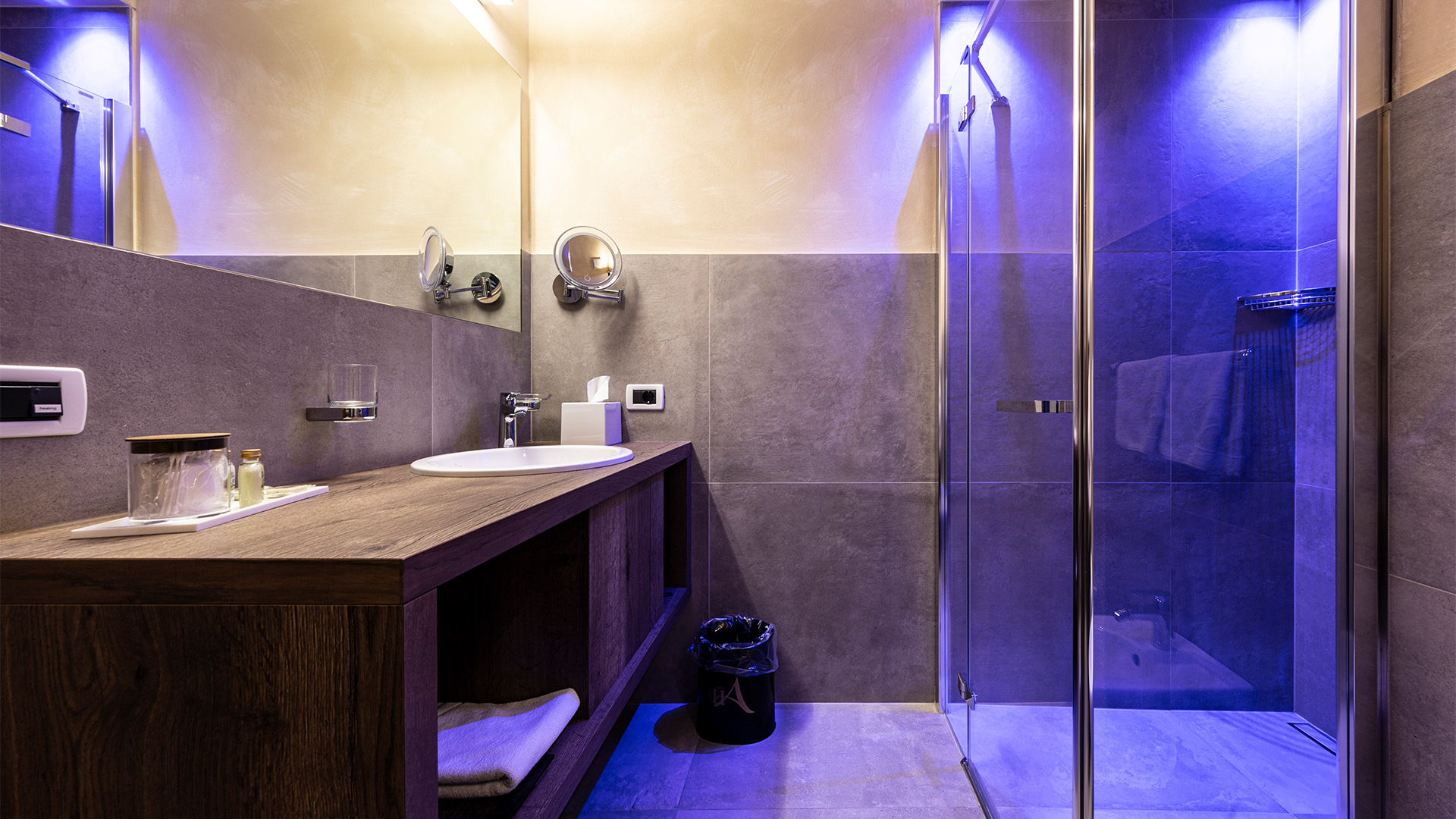 Modernes Badezimmer im Hotel Arkadia mit sanfter blauer Beleuchtung, großem Spiegel und Fliesen in Steinoptik