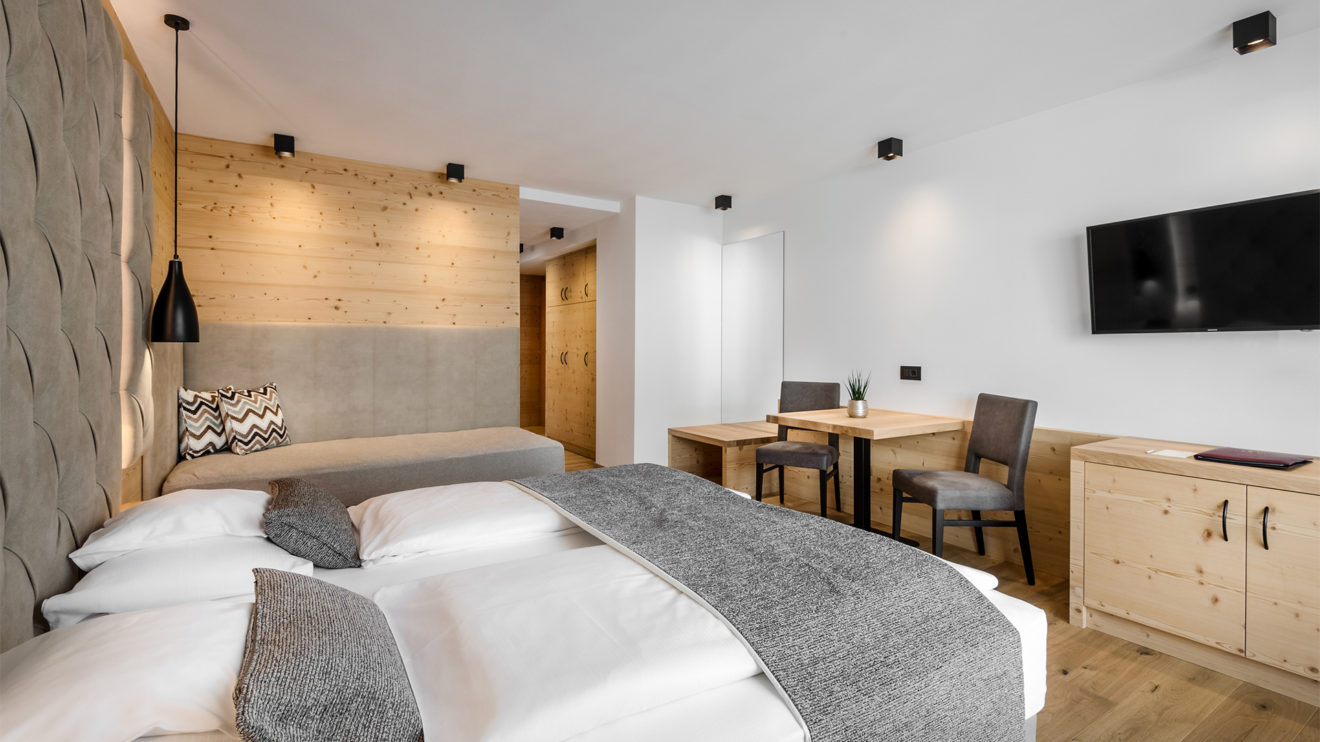 Panorama-Zimmer mit Doppelbett, Wohnbereich mit Sofa, Tisch mit Stühlen, Wand-TV und hellen Holzmöbeln im alpinen Stil
