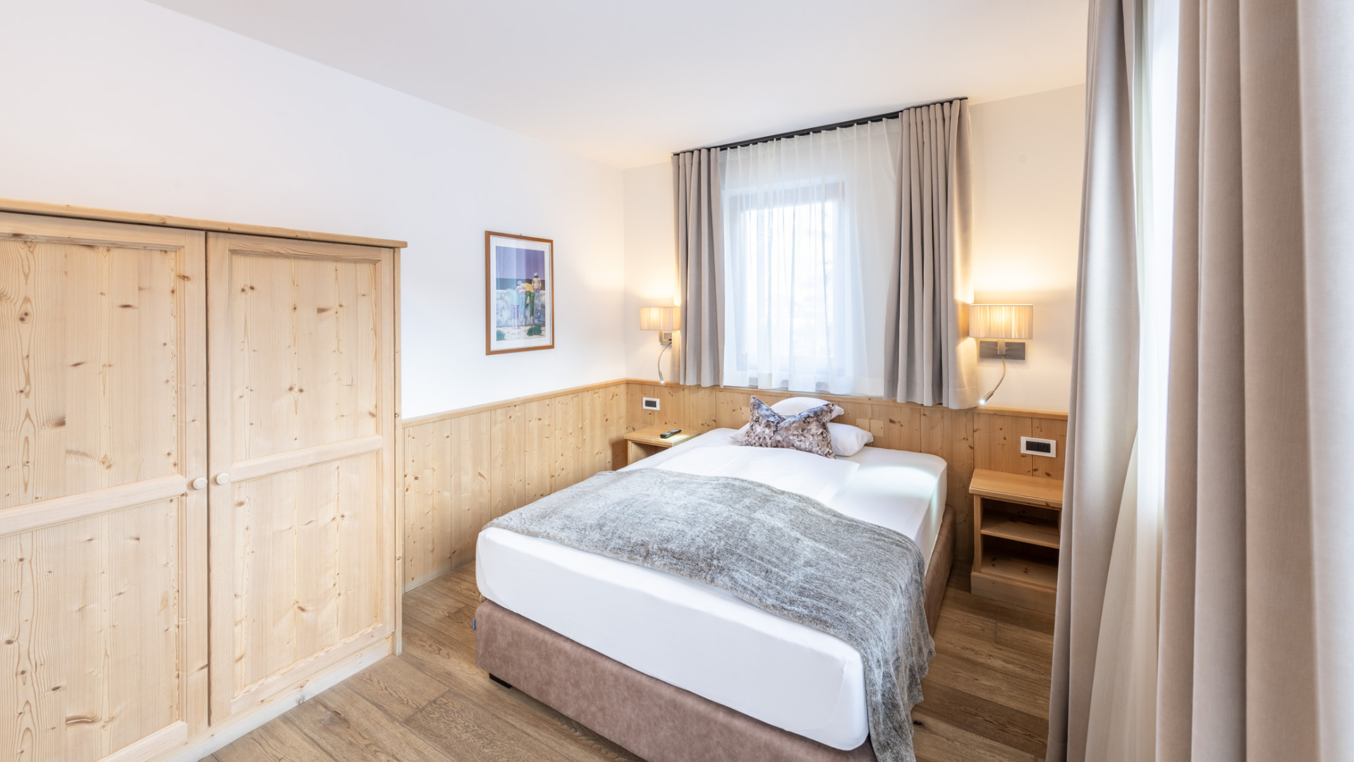 Einzelzimmer im alpinen Stil mit Doppelbett, hellem Holzschrank, Nachttischen mit Leselampen und Fenster mit Vorhängen