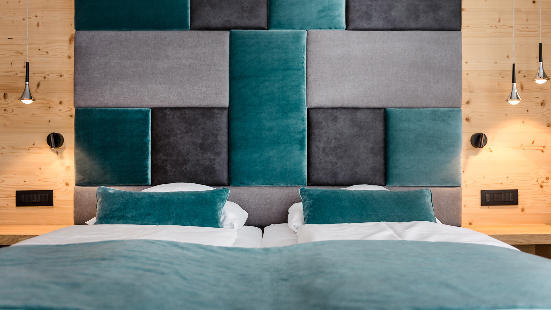 Doppelzimmer mit geometrisch gepolstertem Kopfteil in Petrol- und Grautönen, Doppelbett mit passenden Kissen und Pendelleuchte an einer Holzwand.