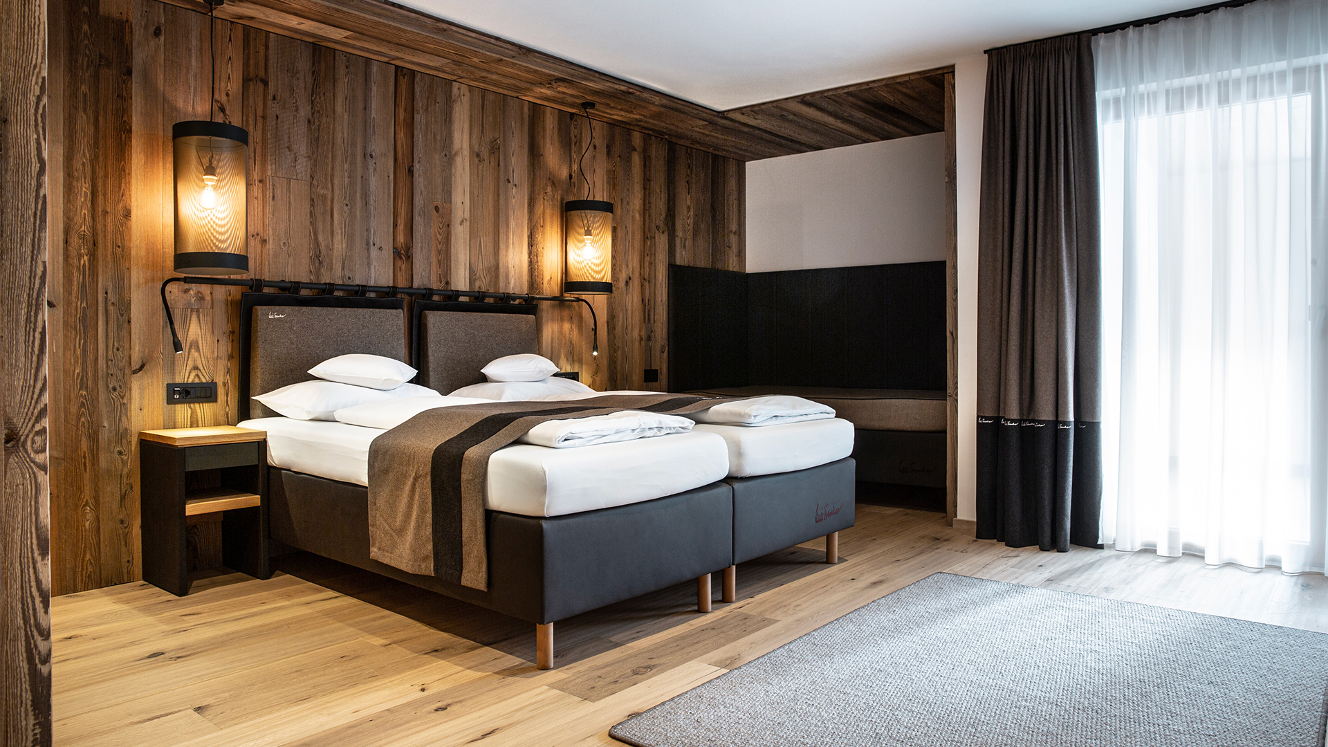 Alpines Hotelzimmer mit Holzwänden, zusammenstehenden Einzelbetten, Hängelampen und einem Entspannungsbereich
