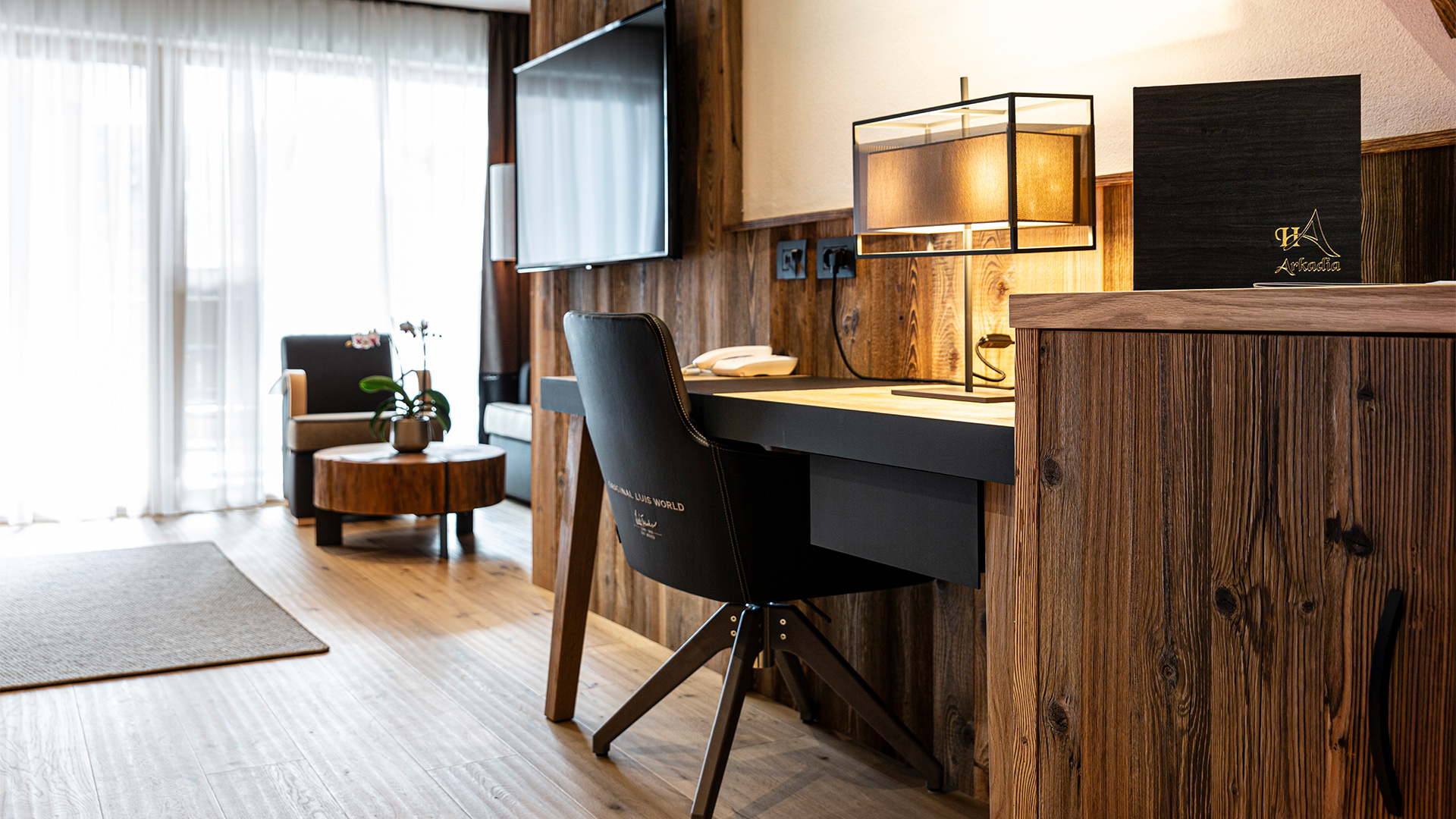 Helles Trenker Zimmer mit Holzschreibtisch, Tischlampe, Wand-TV und Entspannungsbereich mit Sessel und Beistelltisch